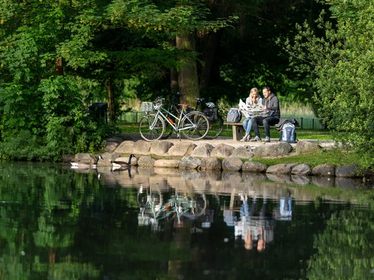 Guetersloh-Stadtpark-Teutoburger-Wald-Tourismus-Patrick-Gawandtka-007.jpg Ein Paar sitzt auf einer Parkbank am Seeufer, Fahrräder stehen daneben, umgeben von üppigem Grün.