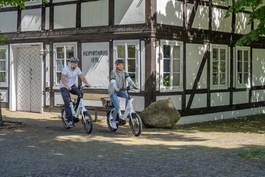 Verl-Heimathaus-Teutoburger-Wald-Tourismus-Patrick-Gawandtka-085.jpg Zwei Radfahrer auf E-Bikes vor einem historischen Fachwerkhaus mit der Aufschrift "Heimathaus Verl".