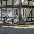 Verl-Heimathaus-Teutoburger-Wald-Tourismus-Patrick-Gawandtka-085.jpg Zwei Radfahrer auf E-Bikes vor einem historischen Fachwerkhaus mit der Aufschrift "Heimathaus Verl".