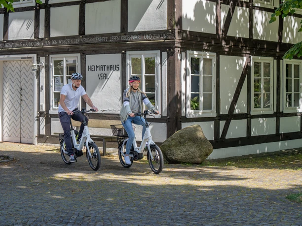 Verl-Heimathaus-Teutoburger-Wald-Tourismus-Patrick-Gawandtka-085.jpg Zwei Radfahrer auf E-Bikes vor einem historischen Fachwerkhaus mit der Aufschrift "Heimathaus Verl".