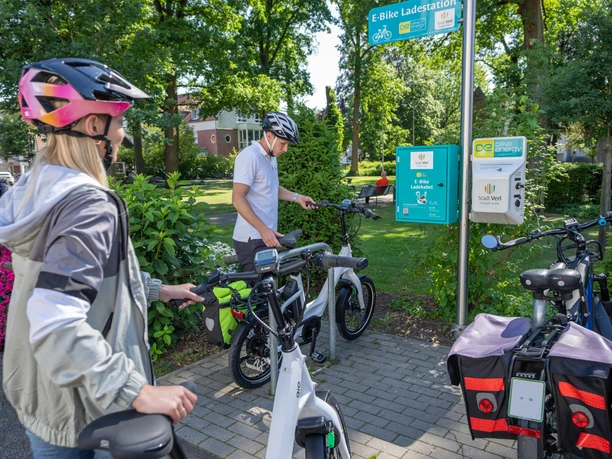 Verl-EBikeLadestation-Teutoburger-Wald-Tourismus-Patrick-Gawandtka-089.jpg Drei Personen pausieren an einer E-Bike-Ladestation im Grünen. Sie tragen Fahrradhelme.