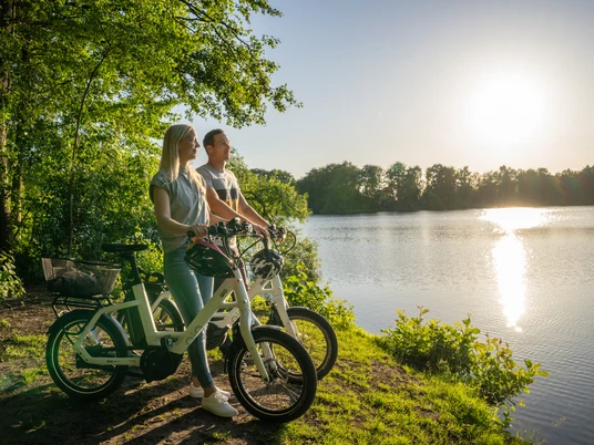 Ein Paar genießt auf E-Bikes den sonnigen Blick über einen ruhigen See, umgeben von Bäumen.