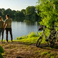 Ein Paar steht am Ufer eines Sees, umgeben von grüner Natur, neben zwei abgestellten Fahrrädern.