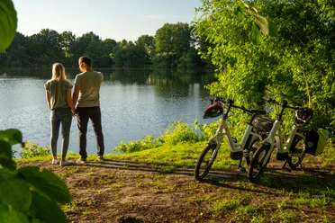 Verl-VerlerSee-Teutoburger-Wald-Tourismus-Patrick-Gawandtka-113.jpg Ein Paar steht am Ufer eines Sees, umgeben von grüner Natur, neben zwei abgestellten Fahrrädern.