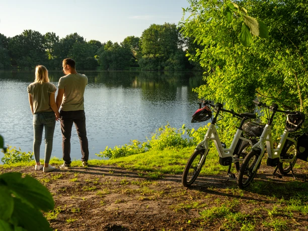 Verl-VerlerSee-Teutoburger-Wald-Tourismus-Patrick-Gawandtka-113.jpg Ein Paar steht am Ufer eines Sees, umgeben von grüner Natur, neben zwei abgestellten Fahrrädern.