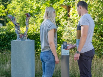 Verl-Skulpturenpark-Teutoburger-Wald-Tourismus-Patrick-Gawandtka-072.jpg Ein Paar betrachtet eine bunte Skulptur im Freien neben einem Infoterminal, umgeben von grüner Natur.