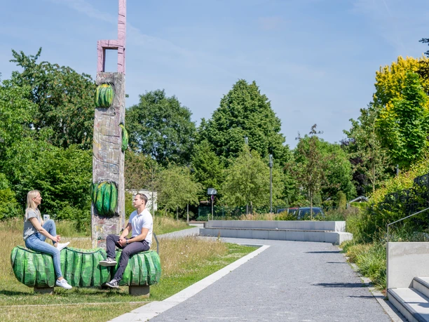 Zwei Personen sitzen auf einer Skulptur im Grünen, umgeben von Bäumen und einem Spazierweg.
