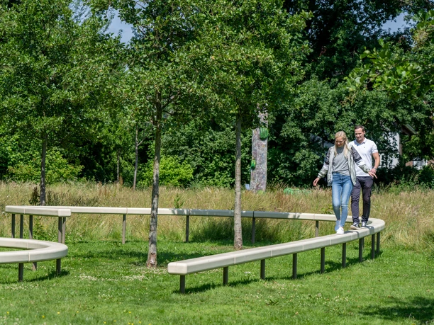 Zwei Personen balancieren fröhlich auf einer geschwungenen Holzbank in einem grünen Park mit Bäumen.