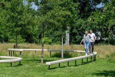 Verl-Skulpturenpark-Teutoburger-Wald-Tourismus-Patrick-Gawandtka-063.jpg Zwei Personen balancieren fröhlich auf einer geschwungenen Holzbank in einem grünen Park mit Bäumen.