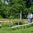 Verl-Skulpturenpark-Teutoburger-Wald-Tourismus-Patrick-Gawandtka-063.jpg Zwei Personen balancieren fröhlich auf einer geschwungenen Holzbank in einem grünen Park mit Bäumen.