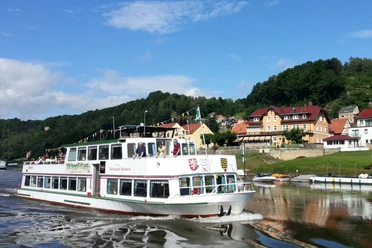 White excursion boat with passengers on a river, in the background green hills and colorful houses under a blue sky.