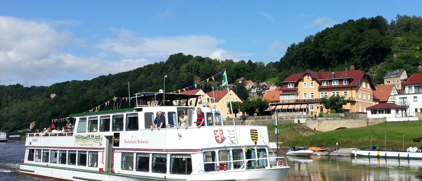 White excursion boat with passengers on a river, in the background green hills and colorful houses under a blue sky.