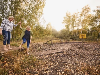 Hille-Torfmoor-Moortreten-Teutoburger-Wald-Tourismus-M-Rothbrust-392.jpg Zwei Frauen überqueren barfuß einen Holzsteg in herbstlicher Landschaft mit Schild daneben.