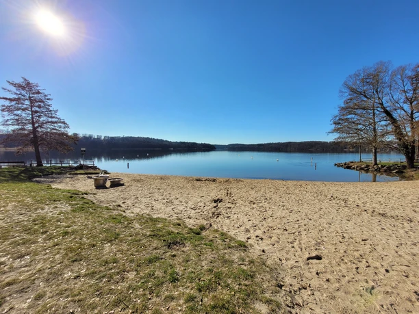 Badestelle_Aqua_Siwa_Silvana Dust_20230228.jpg Sandy lakeshore with calm water, trees on both sides and bright sun overhead
