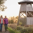 Zwei Frauen wandern auf einem Naturpfad neben einem hölzernen Aussichtsturm bei Sonnenuntergang.