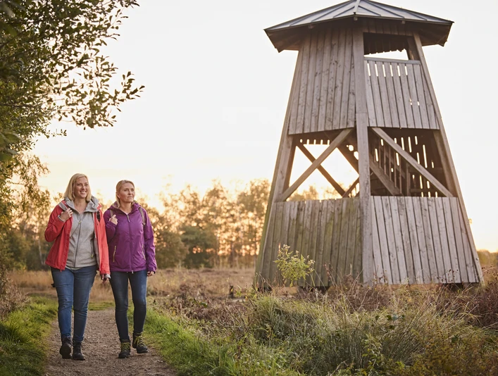 Zwei Frauen wandern auf einem Naturpfad neben einem hölzernen Aussichtsturm bei Sonnenuntergang.