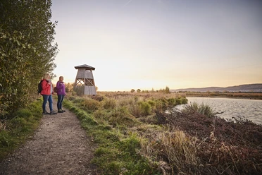 Großes Torfmoor Zwei Frauen wandern bei Sonnenuntergang neben einem ruhigen Flusslauf und einem hölzernen Aussichtsturm.