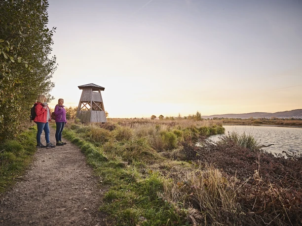 Großes Torfmoor Zwei Frauen wandern bei Sonnenuntergang neben einem ruhigen Flusslauf und einem hölzernen Aussichtsturm.