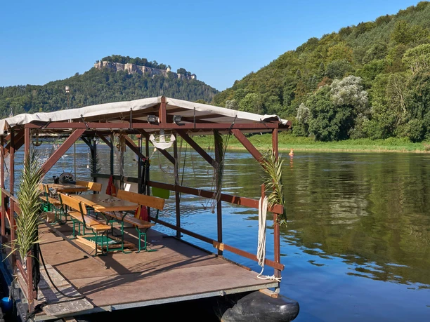 A covered raft with wooden benches and tables floats on a river, surrounded by green hills and a blue sky in the background.