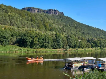 Kanu Aktiv Tours Ein orangefarbenes Motorboot fährt auf einem Fluss, flankiert von grünen Bäumen und einem bewaldeten Hügel im Hintergrund unter klarem, blauem Himmel.