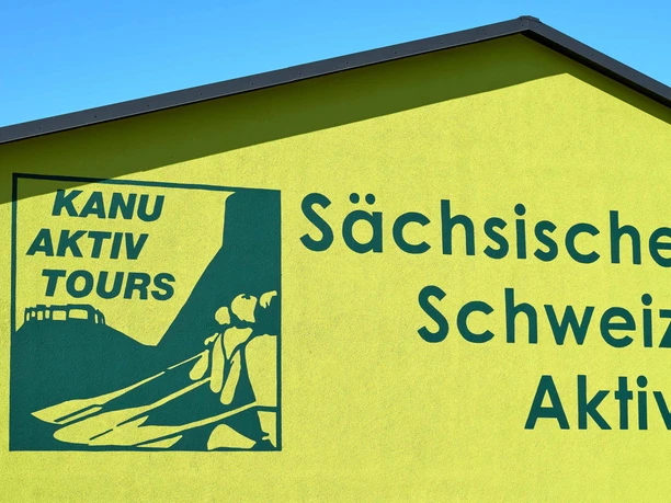 Geel gebouw met blauw logo en opschrift "Kanu Aktiv Tours" en "Sächsische Schweiz Aktiv" onder een blauwe lucht.