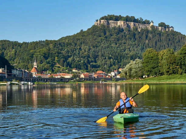 A man paddles in a green kayak on a river, in the background a town with a church and a wooded hill under a clear sky.