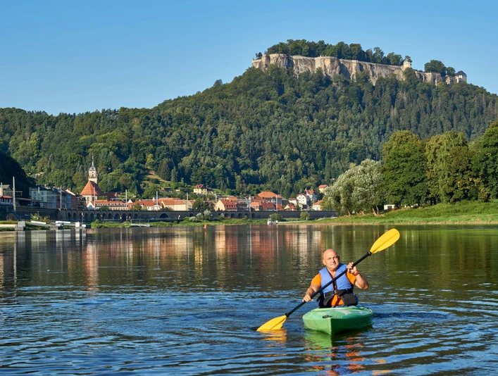Kanu Aktiv Tours Ein Mann paddelt in einem grünen Kajak auf einem Fluss, im Hintergrund eine Stadt mit Kirche und bewaldetem Hügel unter klarem Himmel.