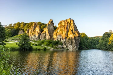Horn-Bad Meinberg-Externsteine-Teutoburger-Wald-Tourismus-D-Ketz-010-CC-BY-SA.jpg Felsenformation in grüner Landschaft spiegelt sich im See, umgeben von Bäumen bei Sonnenuntergang.