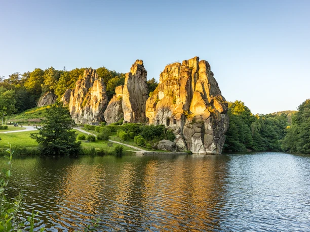 Horn-Bad Meinberg-Externsteine-Teutoburger-Wald-Tourismus-D-Ketz-010-CC-BY-SA.jpg Felsenformation in grüner Landschaft spiegelt sich im See, umgeben von Bäumen bei Sonnenuntergang.