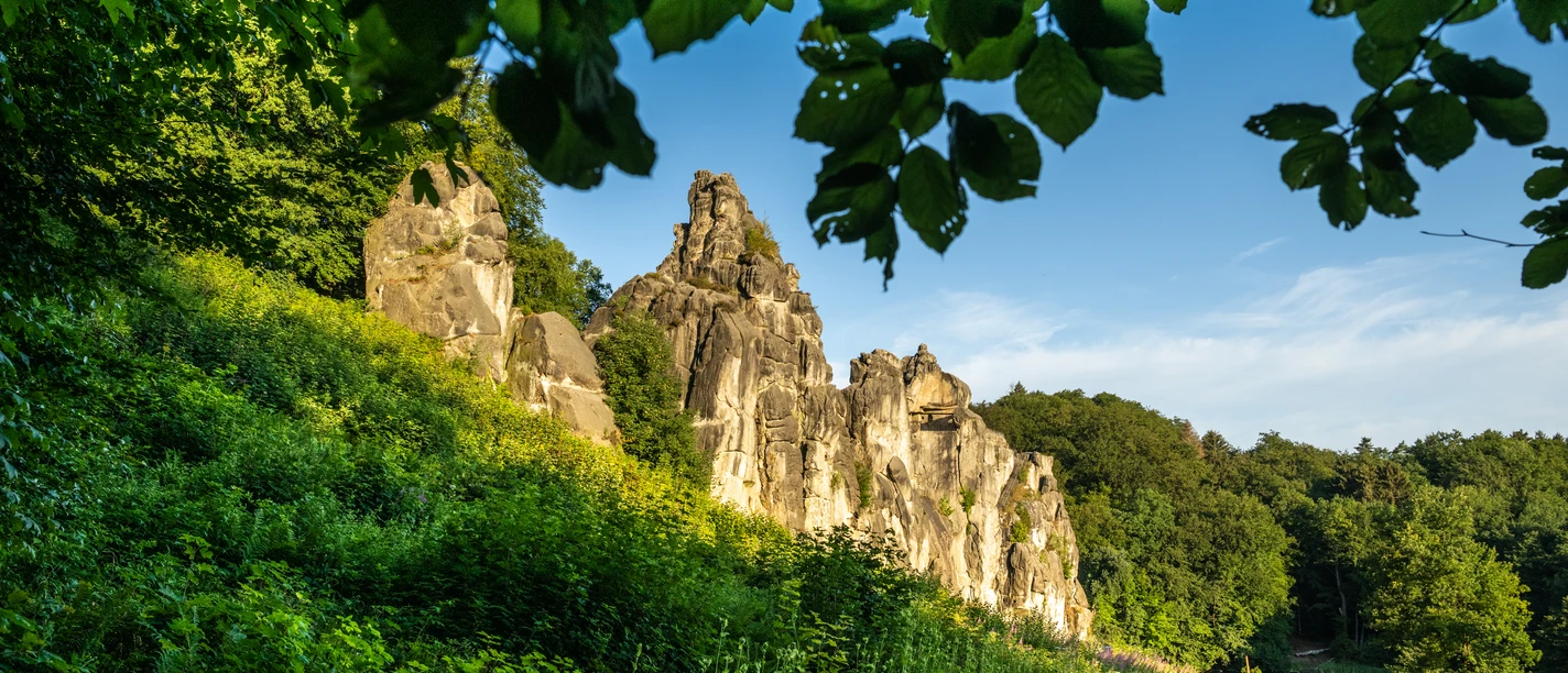 Horn-Bad Meinberg-Externsteine-Teutoburger-Wald-Tourismus-D-Ketz-022-CC-BY-SA.jpg Felsformationen in einer grünen Waldlandschaft, die von einem Kiesweg und Holzzaun gesäumt wird.