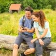 Ein lächelndes Paar in Wanderausrüstung sitzt auf einem Baumstamm und schaut gemeinsam auf ein Handy.