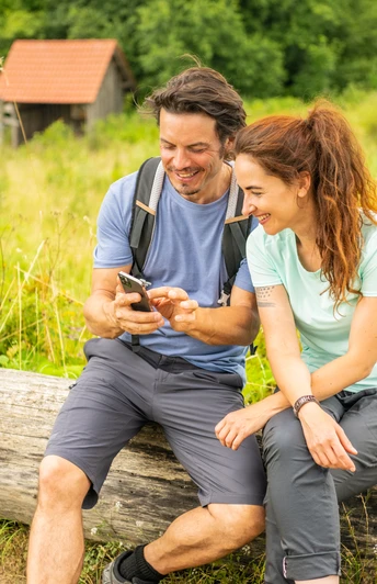 Ein lächelndes Paar in Wanderausrüstung sitzt auf einem Baumstamm und schaut gemeinsam auf ein Handy.