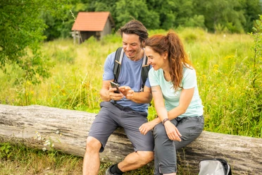 Horn-Bad Meinberg-Preußischer Velmerstot-Teutoburger-Wald-Tourismus-D-Ketz-088.jpg Ein lächelndes Paar in Wanderausrüstung sitzt auf einem Baumstamm und schaut gemeinsam auf ein Handy.