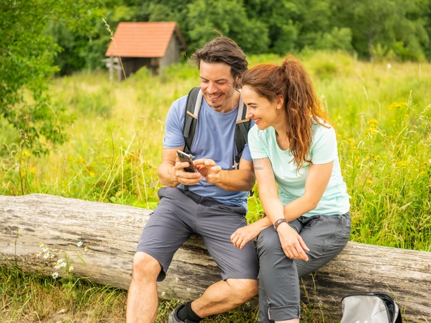 Horn-Bad Meinberg-Preußischer Velmerstot-Teutoburger-Wald-Tourismus-D-Ketz-088.jpg Ein lächelndes Paar in Wanderausrüstung sitzt auf einem Baumstamm und schaut gemeinsam auf ein Handy.