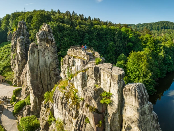 Zwei Personen auf den markanten Felsen der Externsteine, umgeben von üppigem Wald und blauem Himmel.