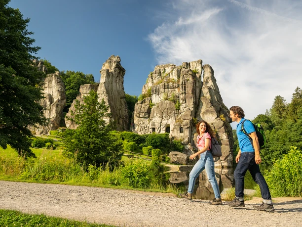 Horn-Bad Meinberg-Externsteine-Teutoburger-Wald-Tourismus-D-Ketz-048.jpg Zwei Wanderer spazieren bei Sonnenschein vor beeindruckenden Sandsteinfelsen in der Natur.