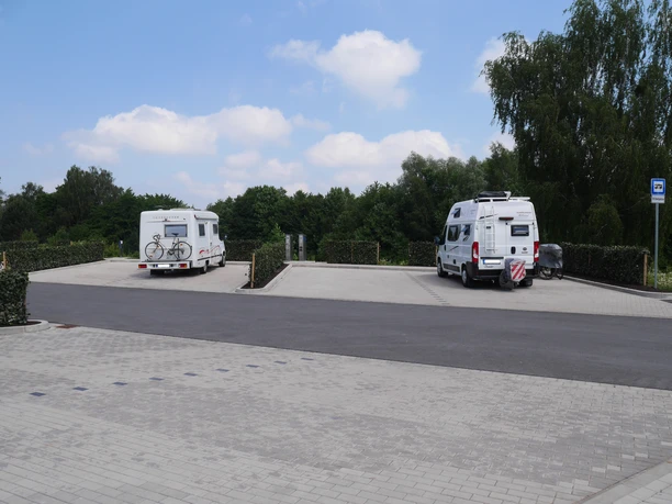 Zwei Wohnmobile parken auf einem gepflasterten Stellplatz, umgeben von grünen Bäumen und Hecken.