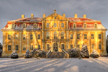 Yellow baroque castle with red roof, lightly covered with snow, in the sunlight; snow-covered trees and bushes in the foreground.