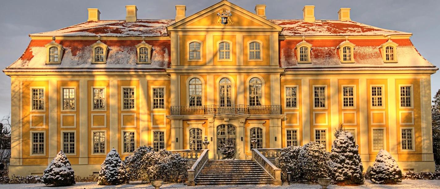 Barockschloss Rammenau Gelbes Barockschloss mit rotem Dach, leicht mit Schnee bedeckt, im Sonnenlicht; verschneite Bäume und Sträucher im Vordergrund.