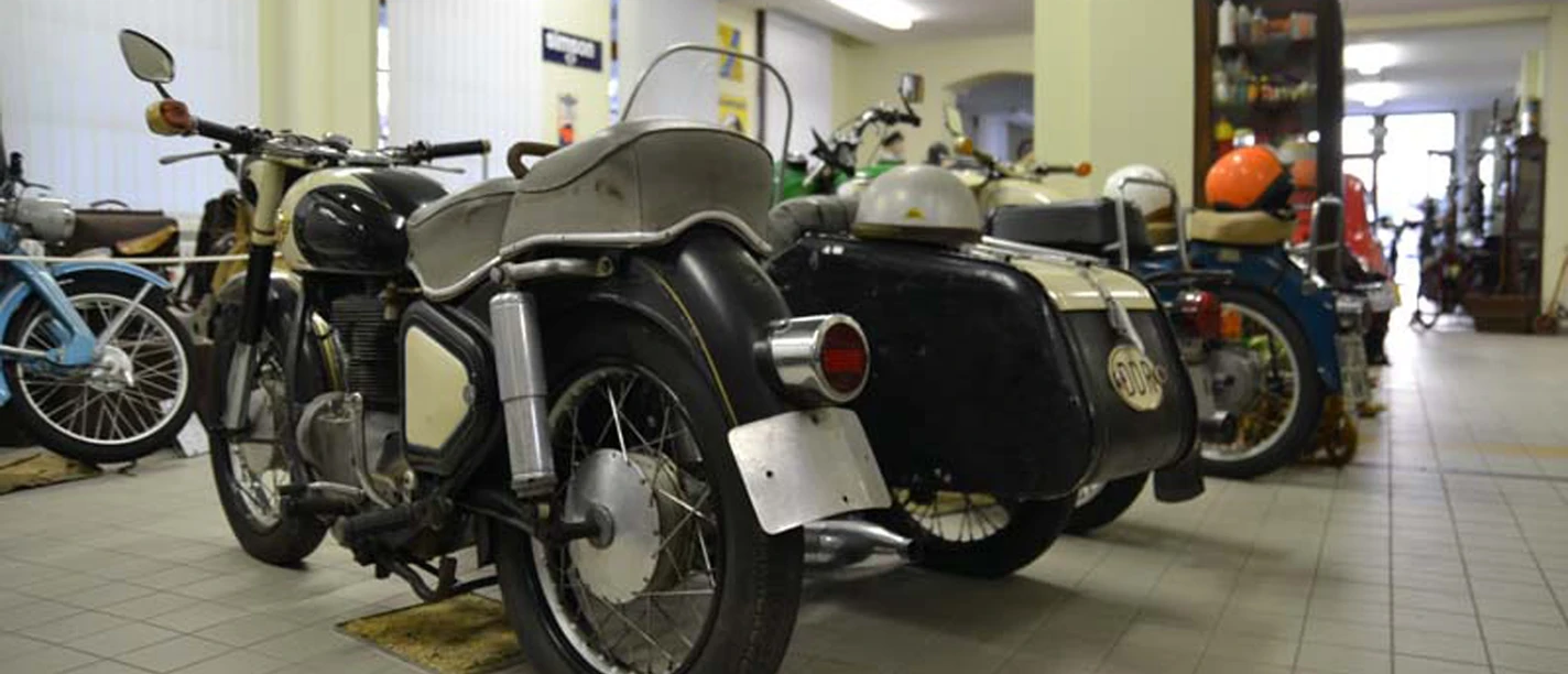 Verschillende klassieke motoren met zijspan staan geparkeerd in een lichte kamer op een tegelvloer, omringd door planken en decor.