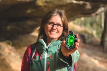 Hiking guide Dr. Anke Dürkopp Woman in outdoor clothing holds a green tag with the inscription "Saxon Switzerland National Park Guide 2022" up to the camera, smiling in the forest.