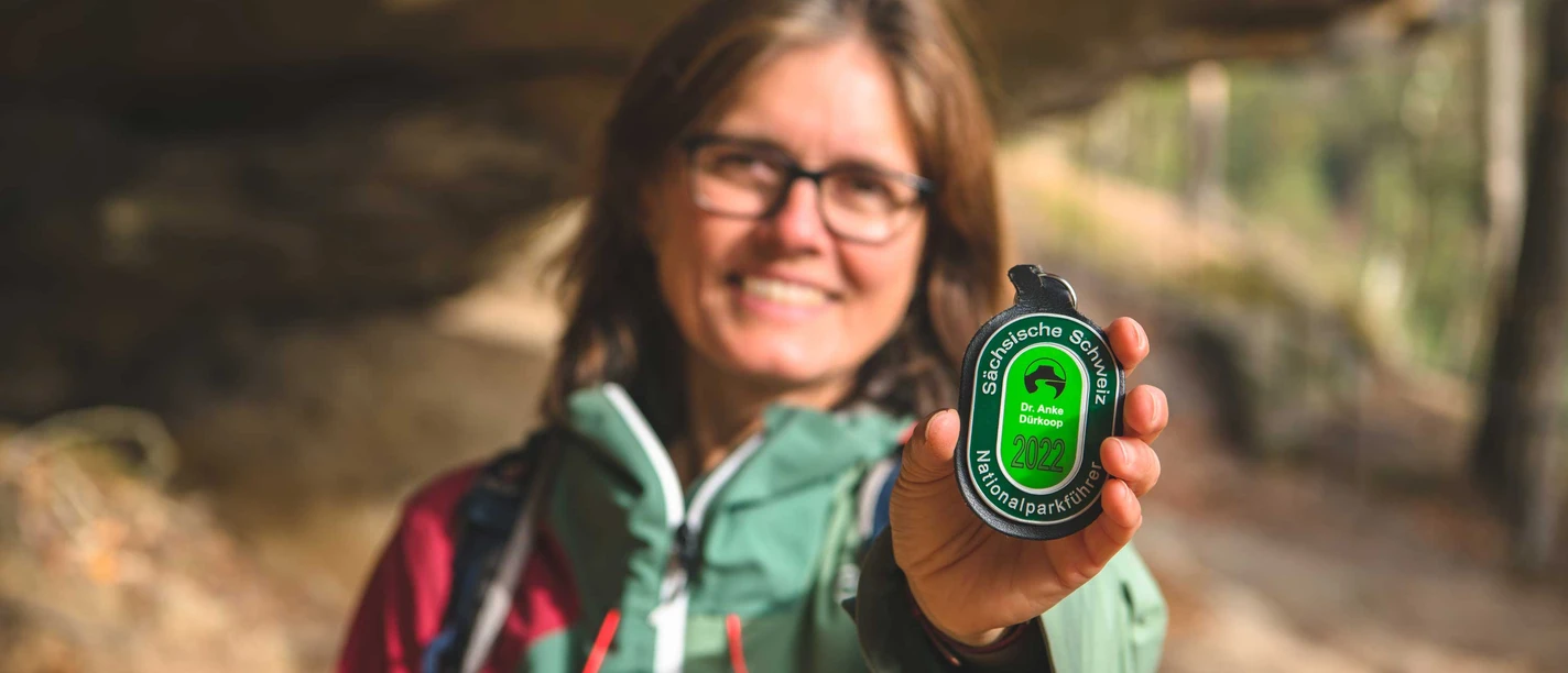 Wanderführerin Dr. Anke Dürkopp Frau in Outdoor-Kleidung hält einen grünen Anhänger mit der Aufschrift "Sächsische Schweiz Nationalparkführer 2022" in die Kamera, lächelnd im Wald.