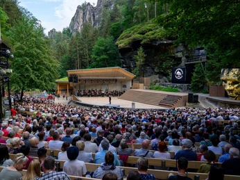 Felsenbühne Rathen Das Freilufttheater namens Felsenbühne Rathen inmitten einer bewaldeten Felslandschaft, gefüllt mit einem großen Publikum, das einer Aufführung auf der Bühne zusieht.The open-air theater called Felsenbühne Rathen in the middle of a wooded rocky landscape, filled with a large audience watching a performance on stage.Divadlo pod širým nebem zvané Felsenbühne Rathen uprostřed zalesněné skalnaté krajiny, zaplněné početným publikem sledujícím představení na jevišti.Teatr na świeżym powietrzu o nazwie Felsenbühne Rathen w środku zalesionego skalistego krajobrazu, wypełniony dużą publicznością oglądającą przedstawienie na scenie.Het openluchttheater genaamd Felsenbühne Rathen midden in een bebost rotslandschap, gevuld met een groot publiek dat kijkt naar een voorstelling op het podium.Il teatro all'aperto chiamato Felsenbühne Rathen, nel mezzo di un paesaggio roccioso e boscoso, è pieno di un grande pubblico che assiste a uno spettacolo sul palco.