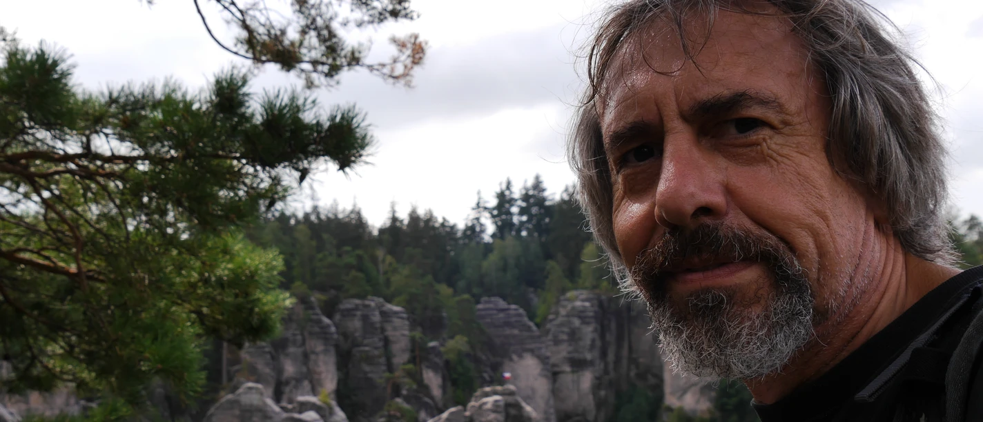 Wanderführer Christian Neumann Ein Mann mit Bart steht vor einer Felslandschaft mit grünen Bäumen und bewölktem Himmel, lächelnd in die Kamera.