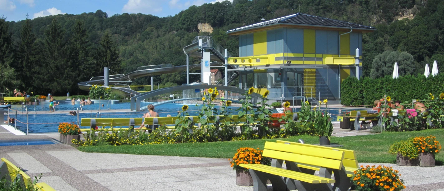Freibad Stadt Wehlen Freibad mit gelben Bänken, Sonnenblumen und einer Wasserrutsche, umgeben von Bäumen und blühenden Pflanzen unter blauem Himmel.