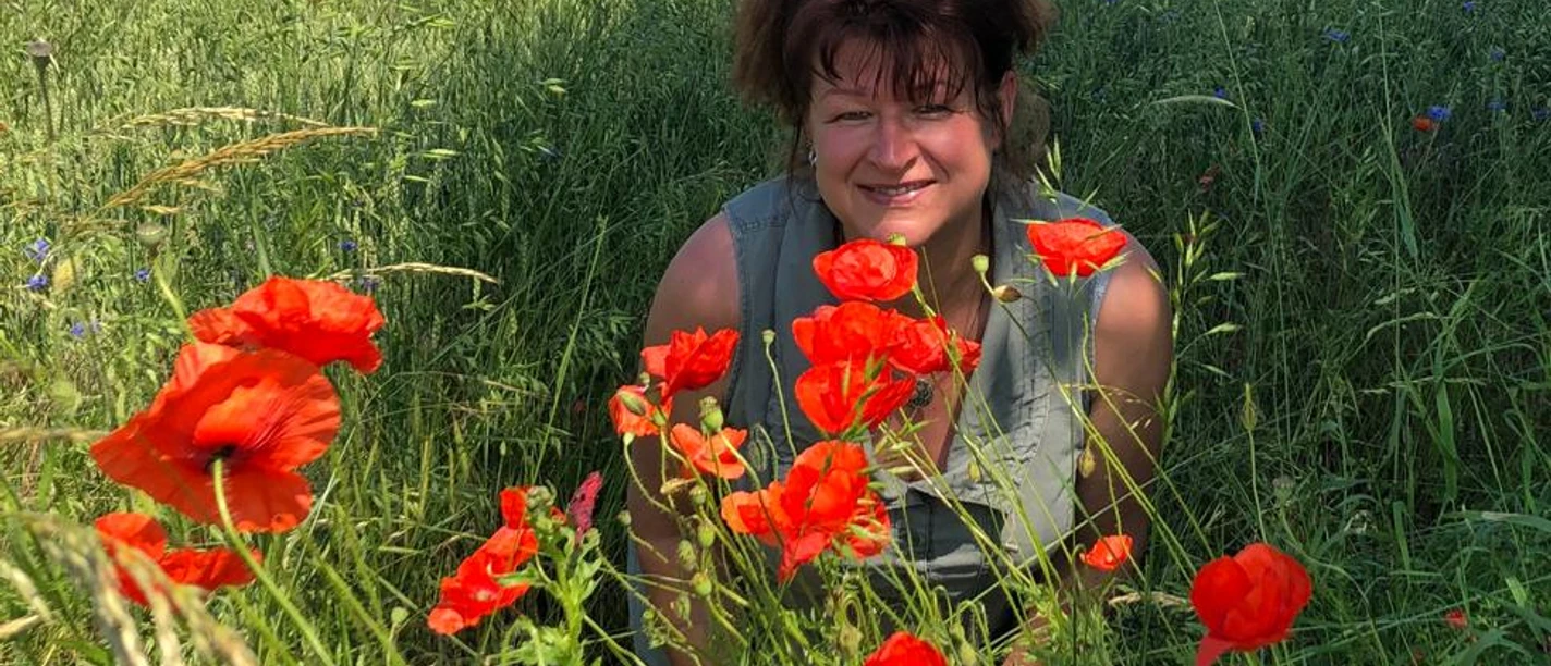 Hiking guide and herbal expert Katrin Vollmann A woman smiles in a green field full of red poppies, with sunny weather and a blue sky in the background.