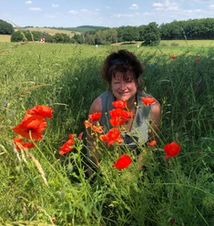 Wanderführerin und Kräuterexpertin Katrin Vollmann Eine Frau lächelt in einem grünen Feld voller roter Mohnblumen, bei sonnigem Wetter und blauem Himmel im Hintergrund.