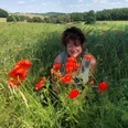 Wanderführerin und Kräuterexpertin Katrin Vollmann Eine Frau lächelt in einem grünen Feld voller roter Mohnblumen, bei sonnigem Wetter und blauem Himmel im Hintergrund.