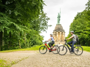 Zwei Radfahrer vor dem Hermannsdenkmal in Detmold, umgeben von üppigem Wald.