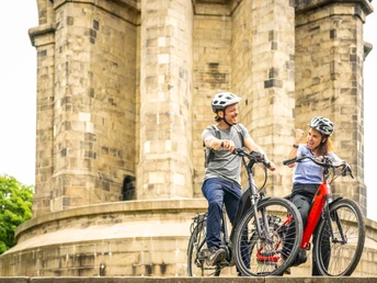 Zwei Radfahrer vor einem historischen Steinturm, lächelnd und in entspannter Unterhaltung.